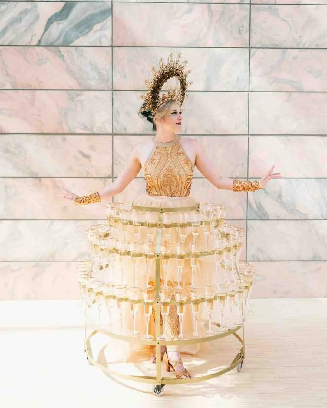 A woman dressed in gold and beige, wearing a large gold headdress and jewelry, standing behind a circular cart with multiple tiers of champagne glasses.