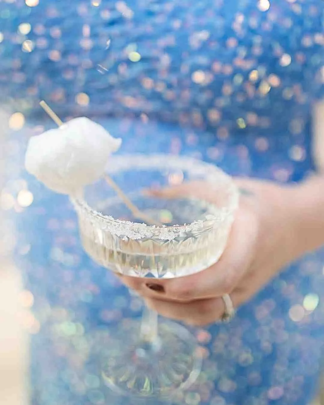 A hand holding a crystal cocktail glass with a salted rim, garnished with cotton candy, set against a sparkling background.