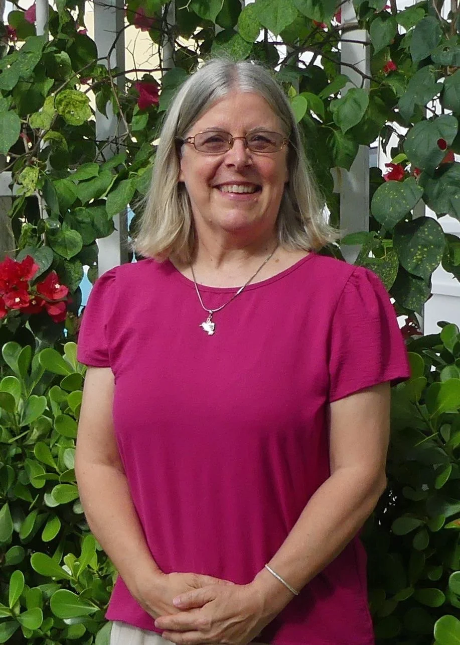 A smiling middle-aged woman with shoulder-length gray hair, wearing glasses, a magenta top, a silver necklace with a bird pendant, and a bracelet, standing in front of green foliage and pink flowers.