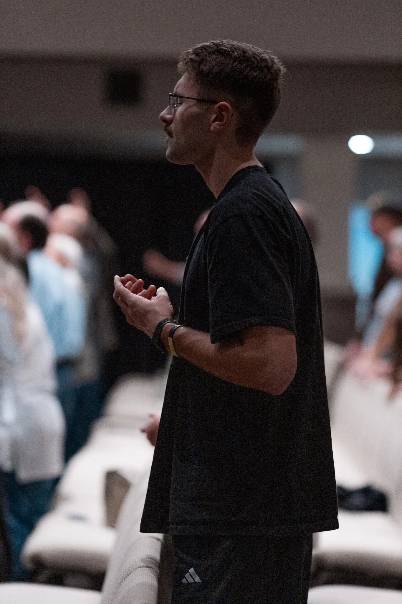A young man with glasses and short hair standing in a darkened room, holding his hands together, with blurred people seated around him.