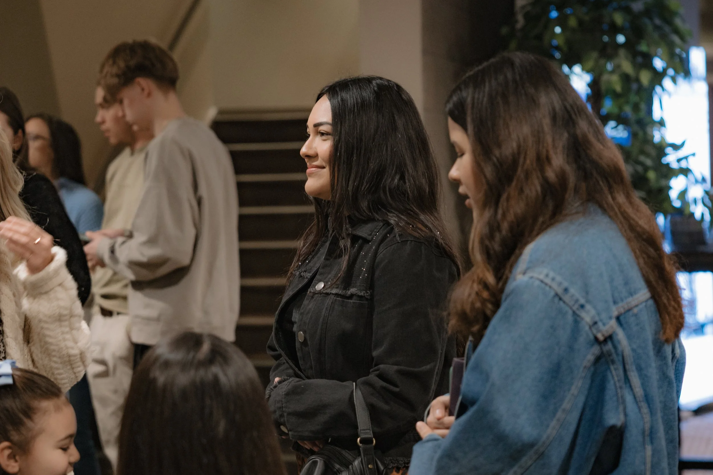 Two women standing in a crowd, one with long black hair in a black jacket smiling, and the other with long wavy brown hair in a blue denim jacket, surrounded by children and people in an indoor setting.
