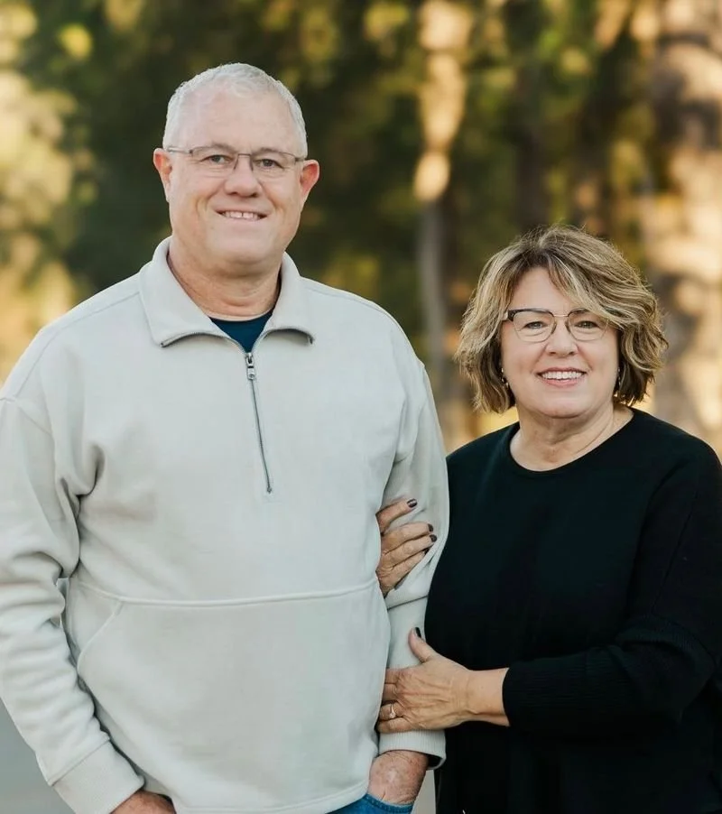 A smiling older couple outdoors, standing close together with greenery in the background.