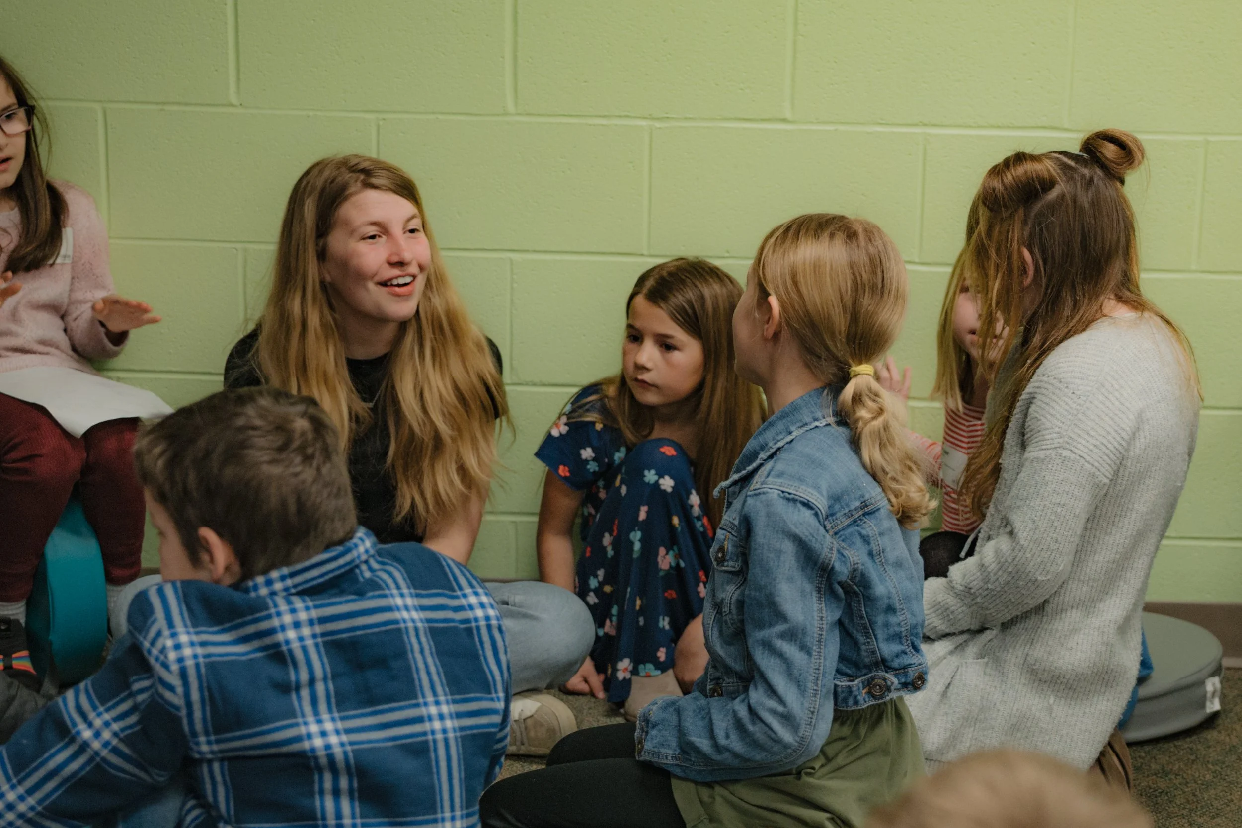 Group of children sitting on the floor against a green brick wall, engaging in conversation.