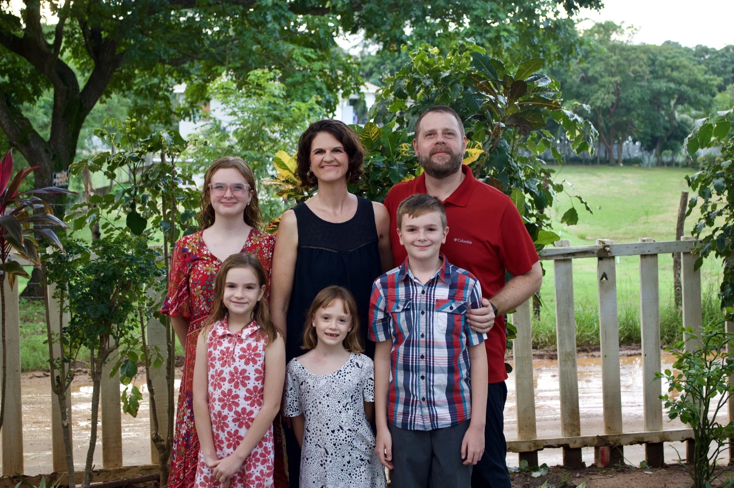 A family of seven standing outdoors in a garden with trees and greenery in the background, smiling at the camera.