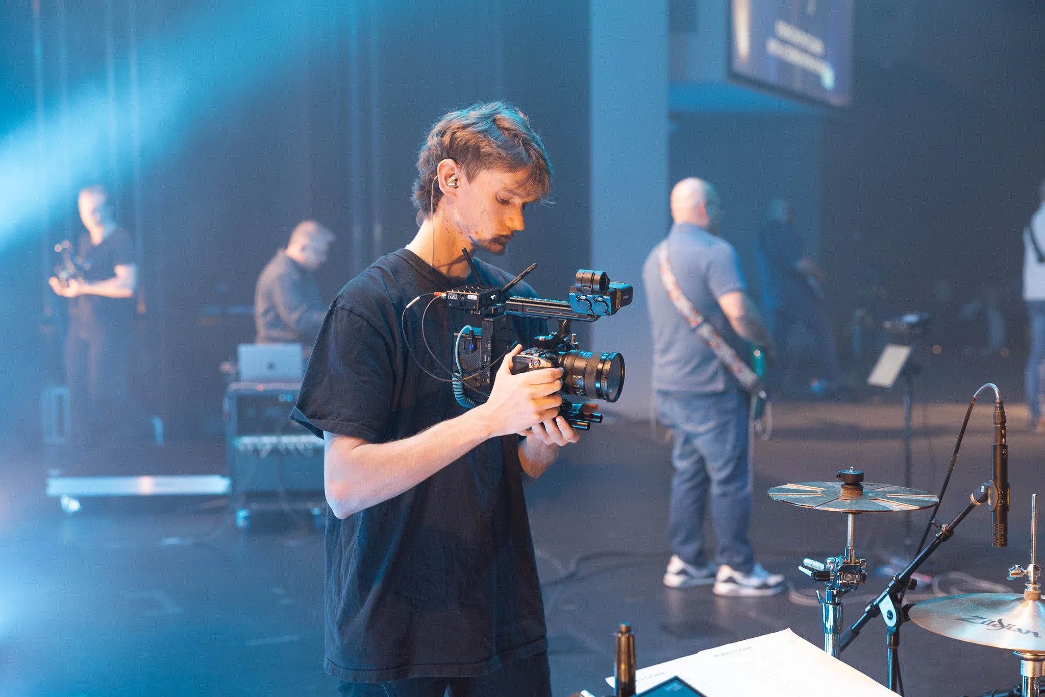 A young man operating a professional video camera on a stage with musicians in the background.