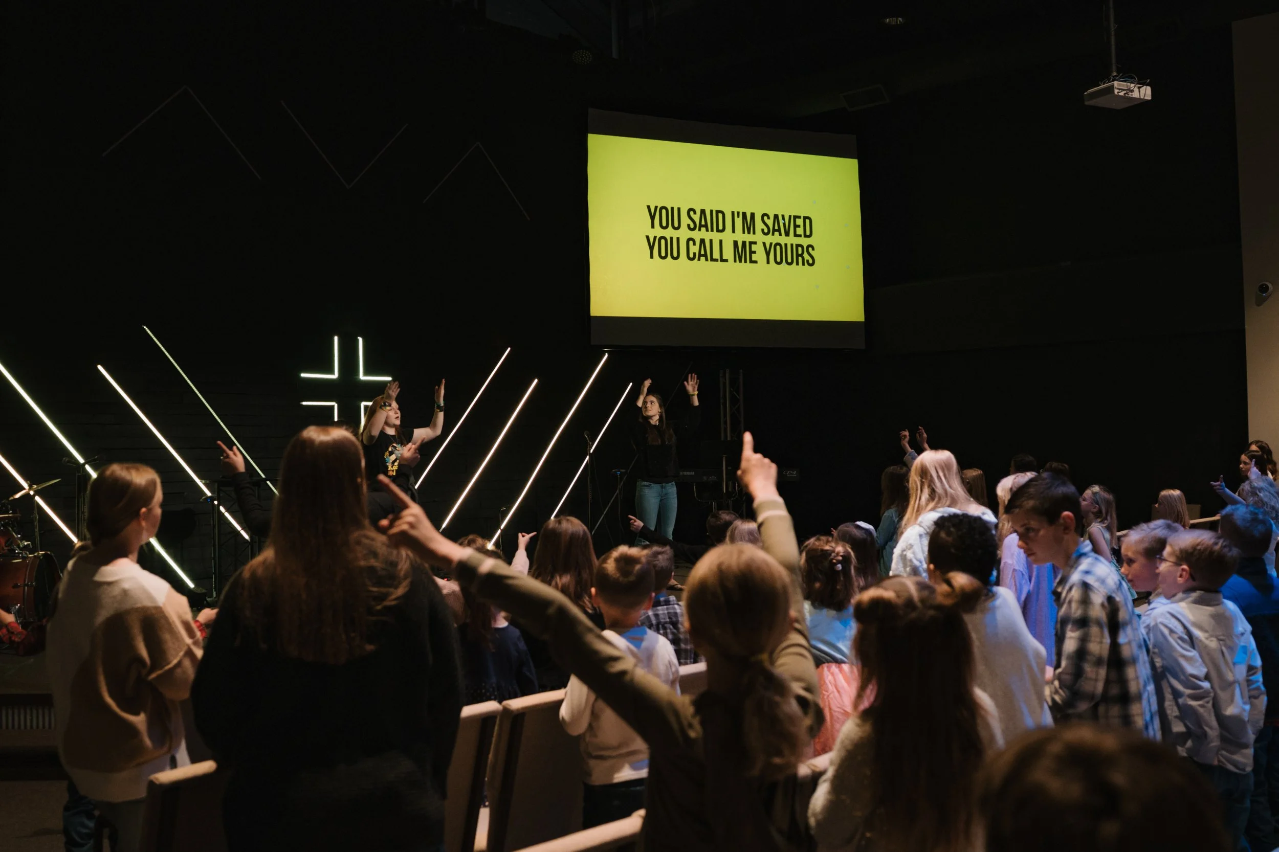 A group of children gathered in a church or concert hall, participating in a worship or singing session, with some children raising their hands. On the stage, two women lead the activity, with a large screen displaying lyrics that say, "You said I'm 