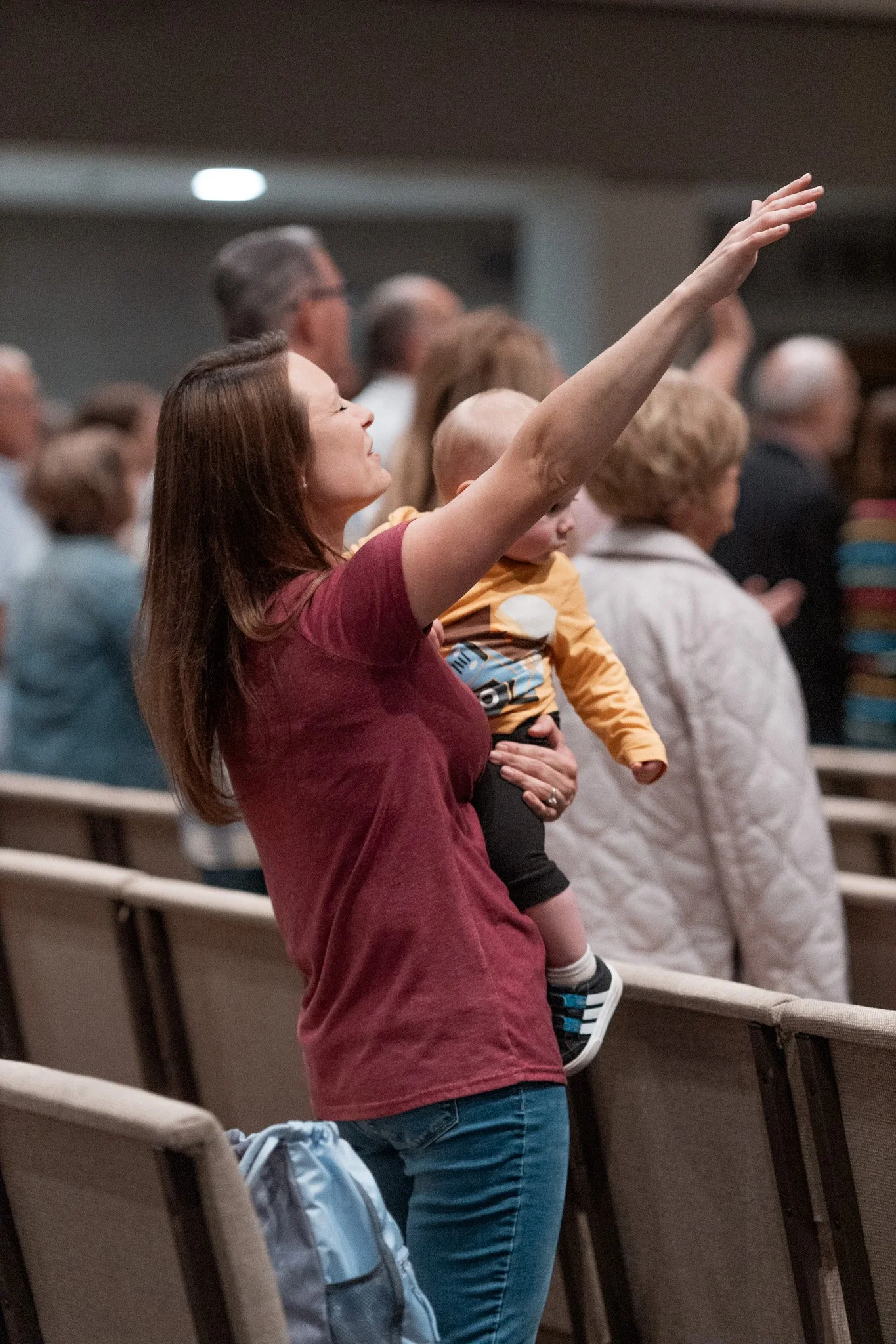 A woman holds a young child and raises her hand in a crowded indoor gathering, possibly a church or conference, with other people standing nearby.
