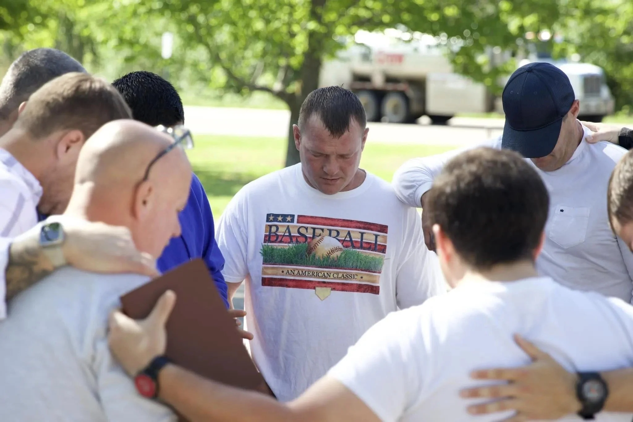 A group of men are gathered outdoors in a circle, with their arms around each other, engaged in a prayer or moment of reflection. One man in the center is wearing a white T-shirt with a baseball graphic and the words 'An American Classic', and the background shows green trees and a parked truck.