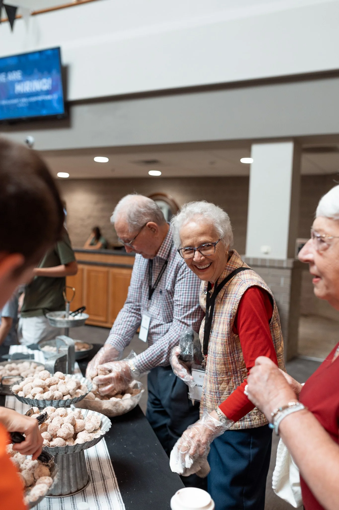 Older adults serving powdered donuts at a food table, smiling and chatting.