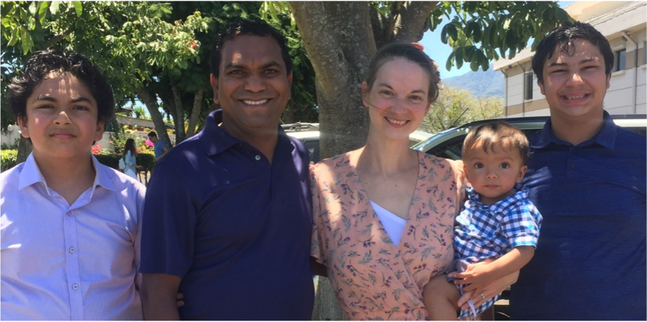 Family of five posing outdoors, standing in front of a tree, with a house and cars in the background. They are smiling, with three children and two adults.