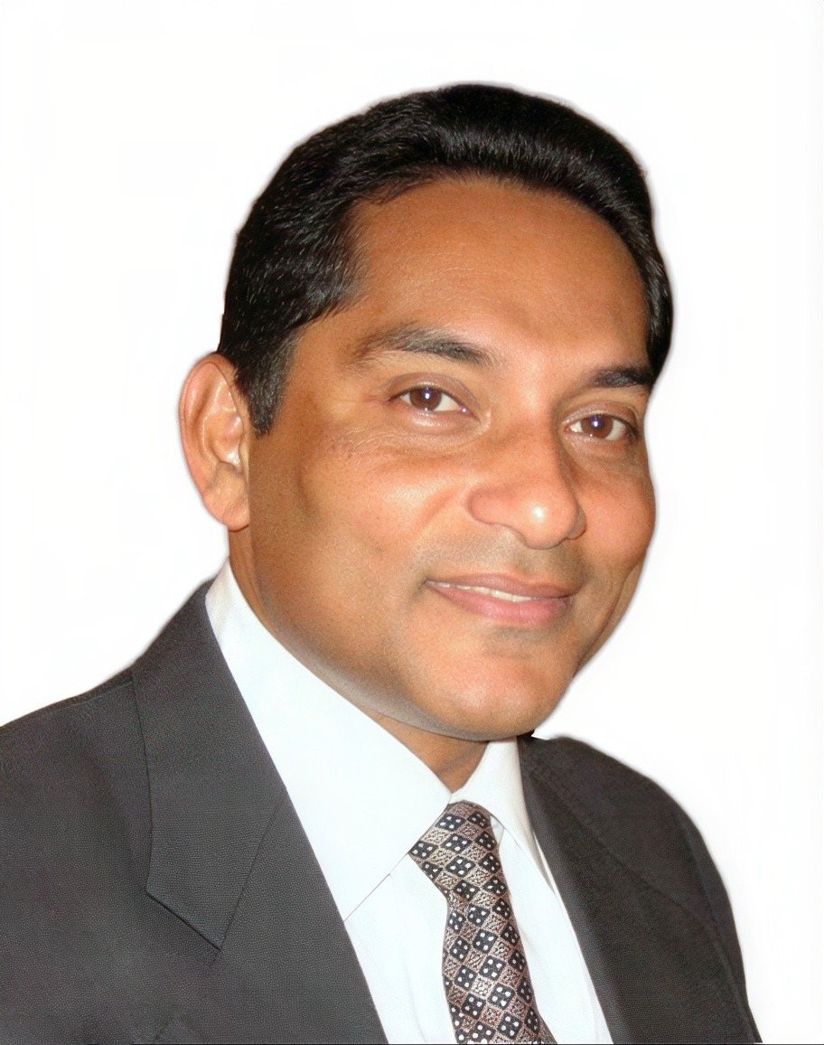 Headshot of a man wearing a suit, white shirt, and patterned tie, smiling against a plain white background.