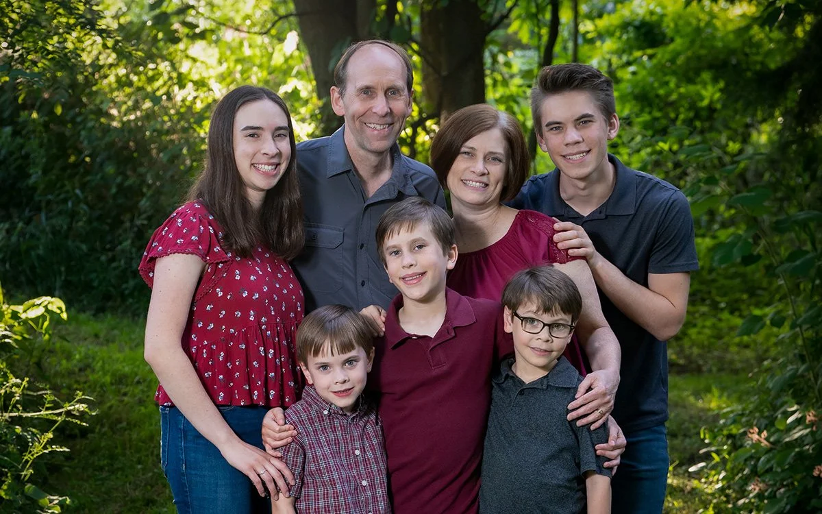 A family of seven, including parents and children, smiling and standing together outdoors in a lush green park.