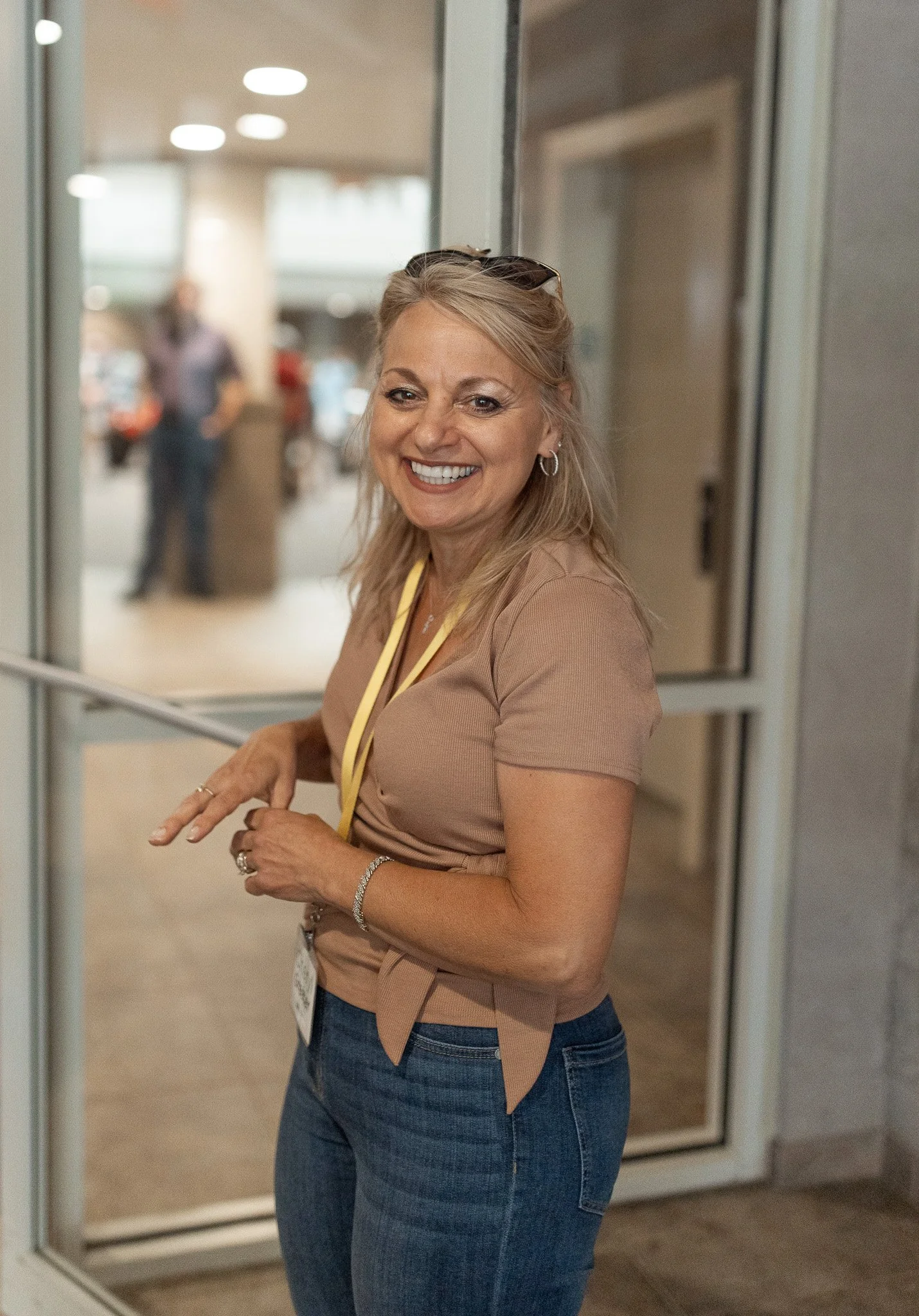 A smiling woman with blonde hair and sunglasses on her head standing near a glass door.