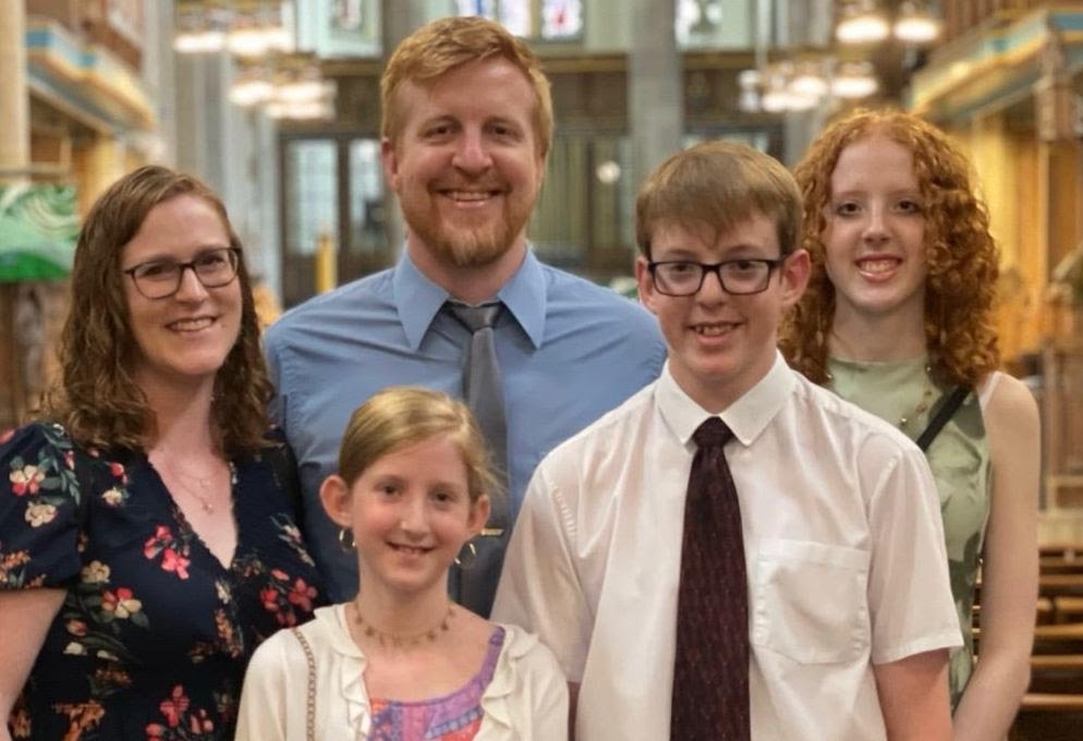 Family of six posing outdoors in front of a house window. Two adults, one man and one woman, along with three children, one girl and two boys, smiling at the camera.