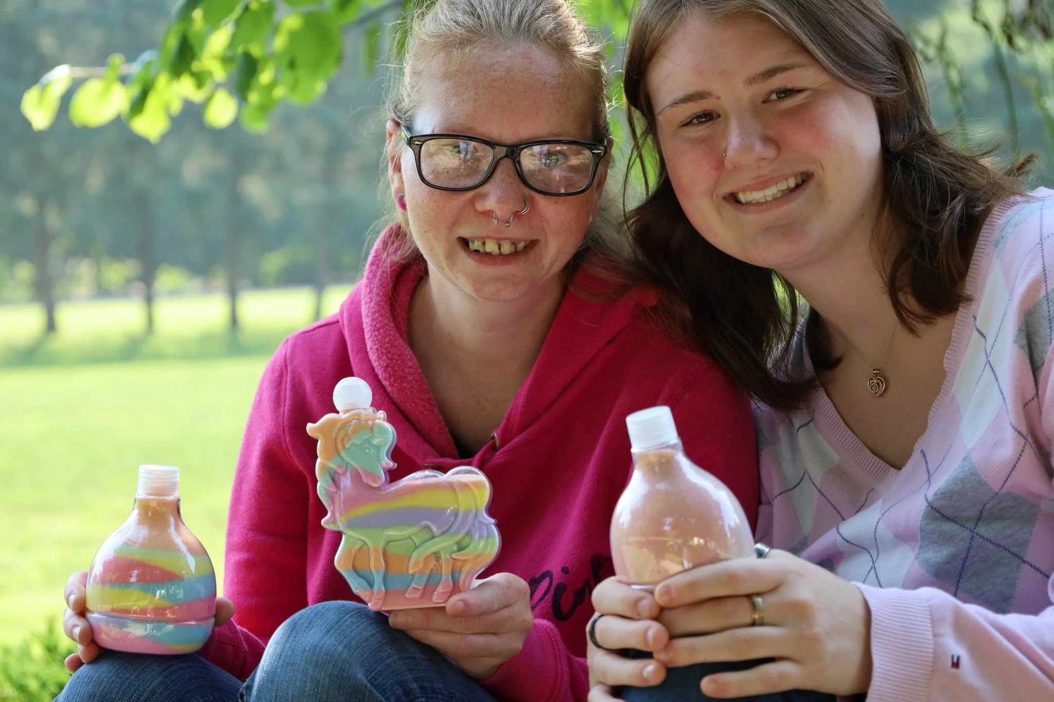 Two young women sitting outdoors on grass, holding bubble bottles and decorated unicorn soap containers, smiling at the camera with a background of trees and greenery.