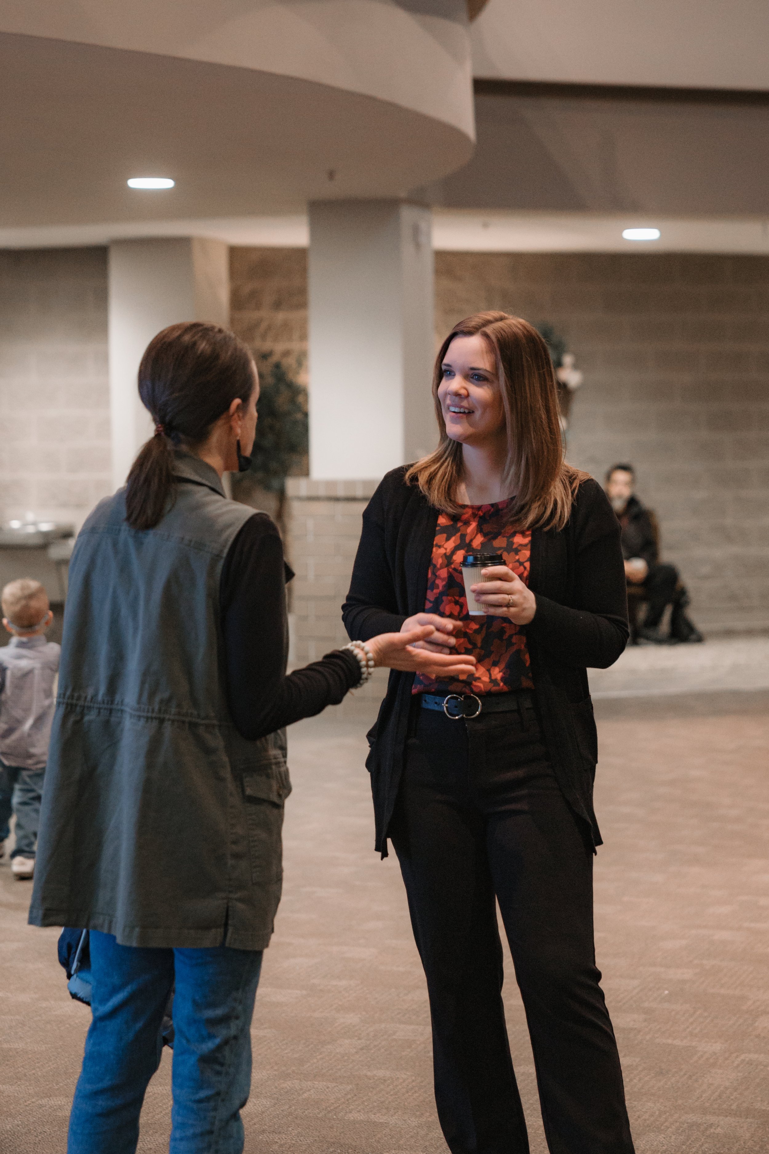 Two women are engaged in a conversation in an indoor setting, with one holding a coffee cup. They are smiling and appear to be enjoying a friendly chat, with a few other people and children visible in the background.