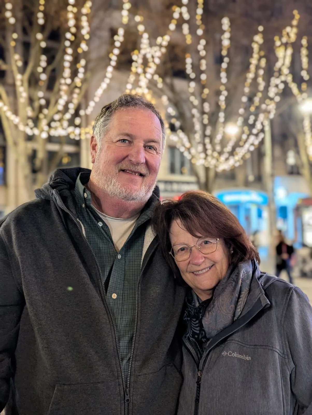 A smiling middle-aged man with a beard and a woman with glasses, both dressed in jackets, standing close together outdoors at night during the winter season with festive string lights above in an urban setting.