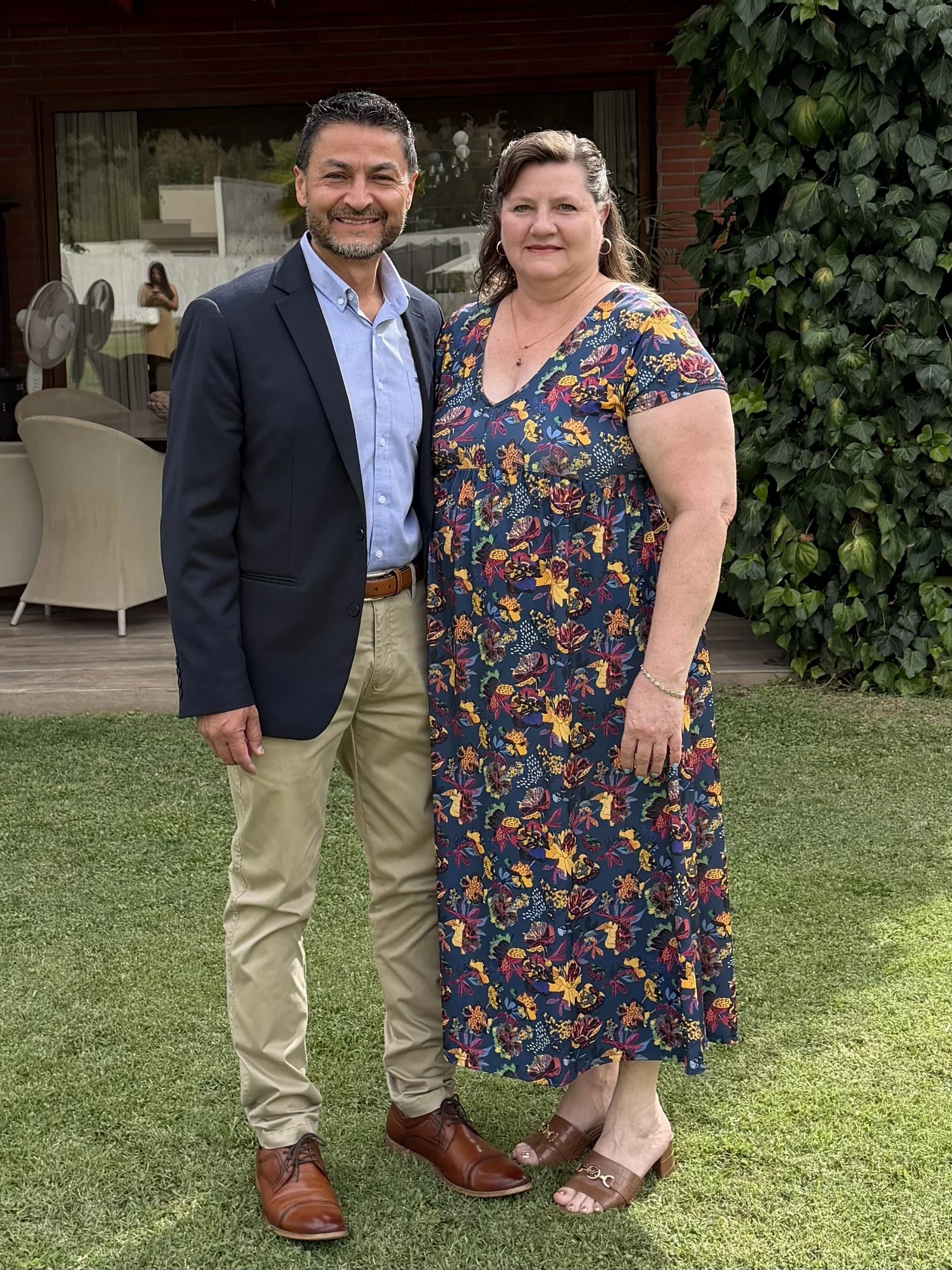 A man and woman stand together outdoors on a lawn, smiling at the camera. The man has dark hair and a beard, is wearing a navy blazer, light blue shirt, beige pants, and brown shoes. The woman has long, wavy brown hair, and is wearing a floral dress with yellow, purple, and red patterns, and brown sandals. Behind them is a house with a large window, plants, and a patio with chairs.