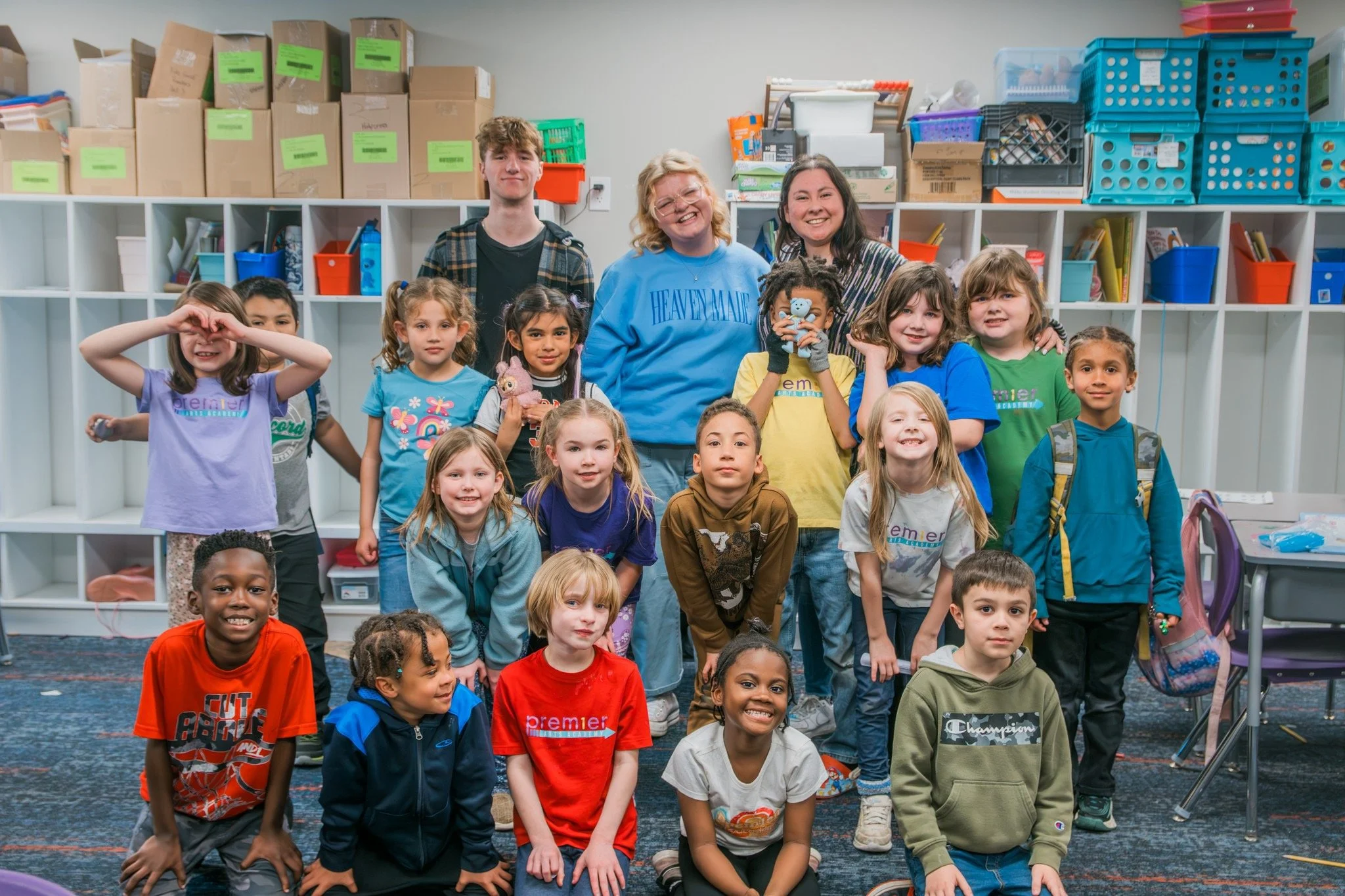 A diverse group of children and three adults posing inside a classroom, with storage shelves and colorful bins in the background.
