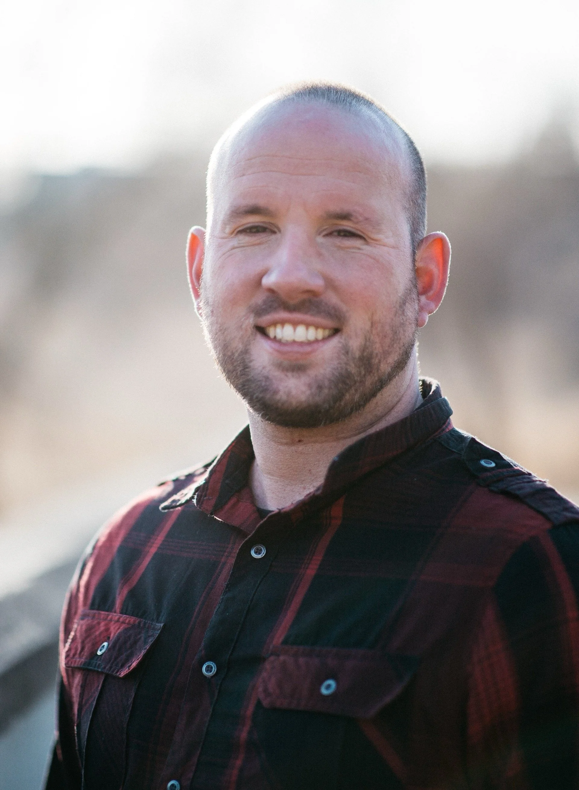 A smiling man with a beard, wearing a black and red plaid shirt, outdoors on a sunny day.