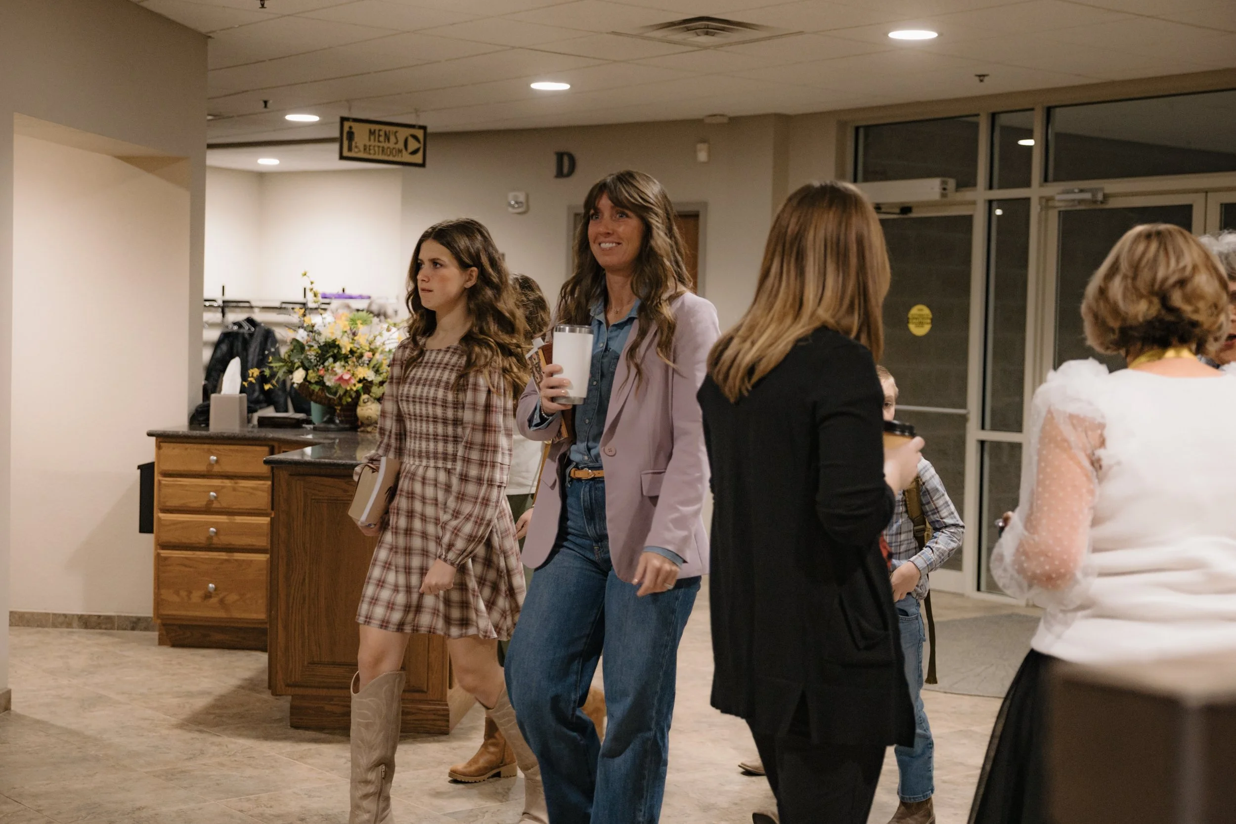 Group of women socializing indoors, one holding a glass of milk, in a casual setting with flowers and coat rack in the background.