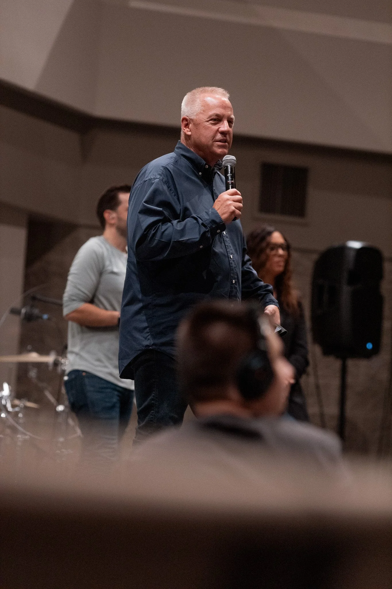 A man with white hair, wearing a dark blue shirt, holding a microphone and speaking at an indoor event. There are two women in the background and a person in the foreground with headphones.