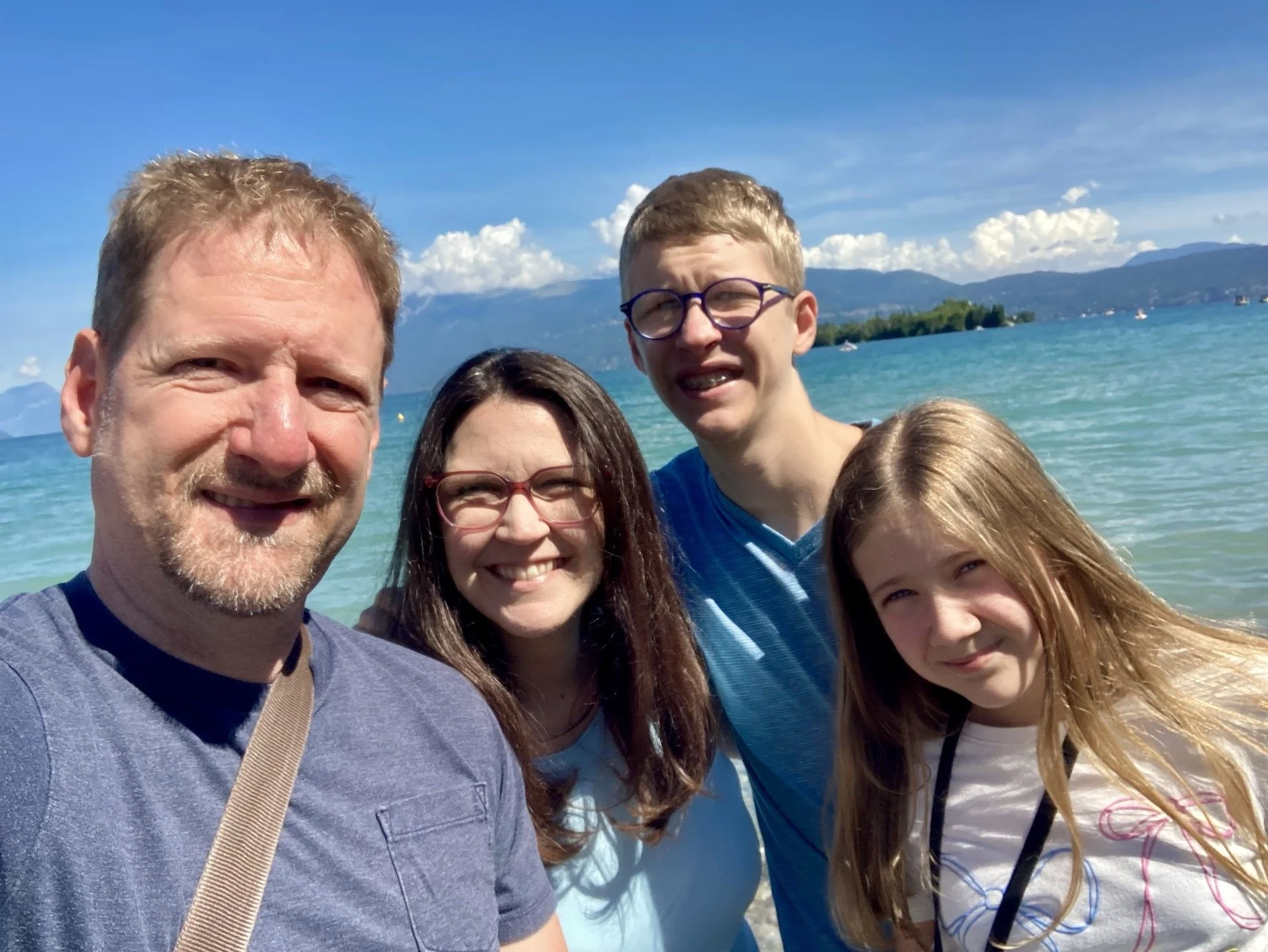 A family of four taking a selfie on a beach with a lake, mountains, and blue sky in the background.