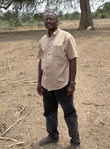 A man standing outdoors on dirt ground, smiling, wearing glasses, a beige short-sleeved shirt, black pants, and a watch, with trees and a small animal in the background.