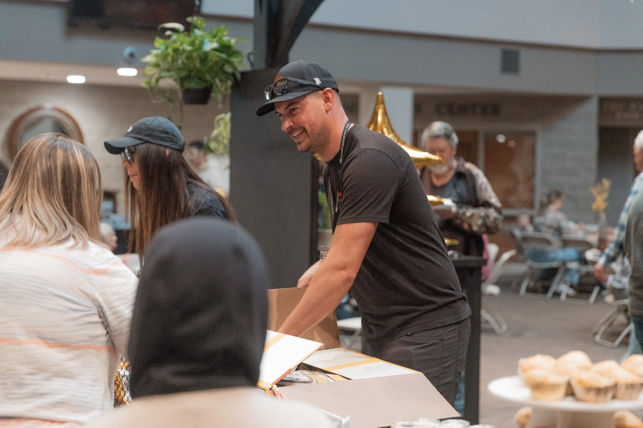 A smiling man in a black t-shirt and black cap handing out food or items at a table indoors, with people and decorations in the background.