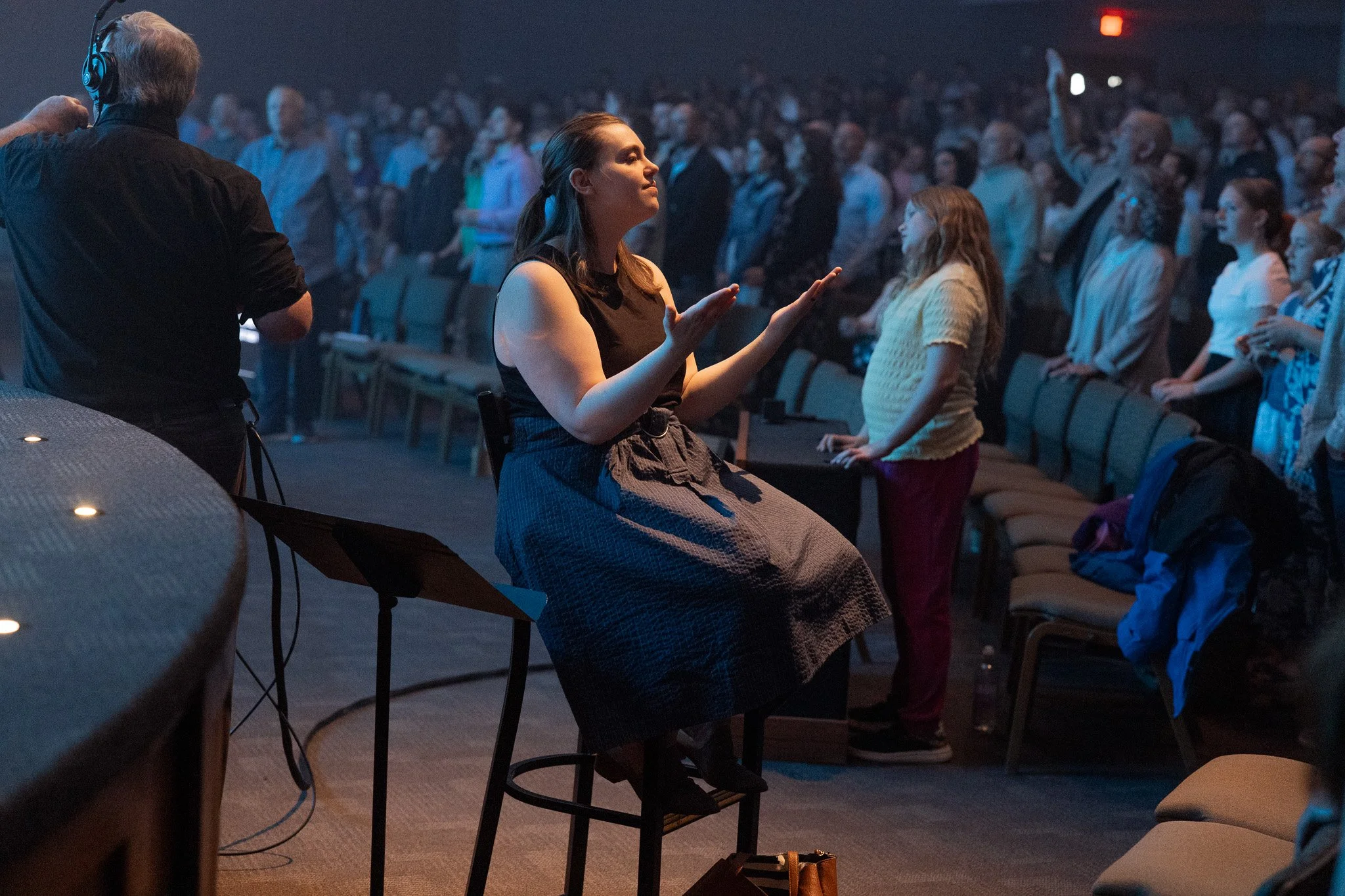A woman is sitting on a stool with her eyes closed, arms raised, and hands open, appearing to be praying or meditating, in a large indoor auditorium filled with a crowd of people standing and facing forward.