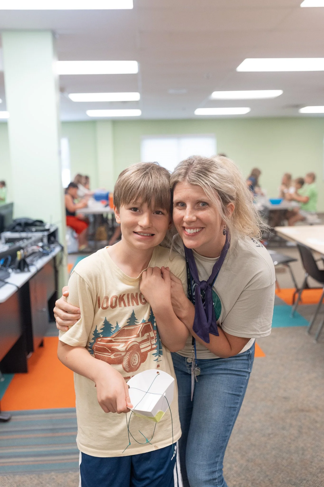 A woman and a boy smiling and hugging in a classroom or camp setting with other children in the background.
