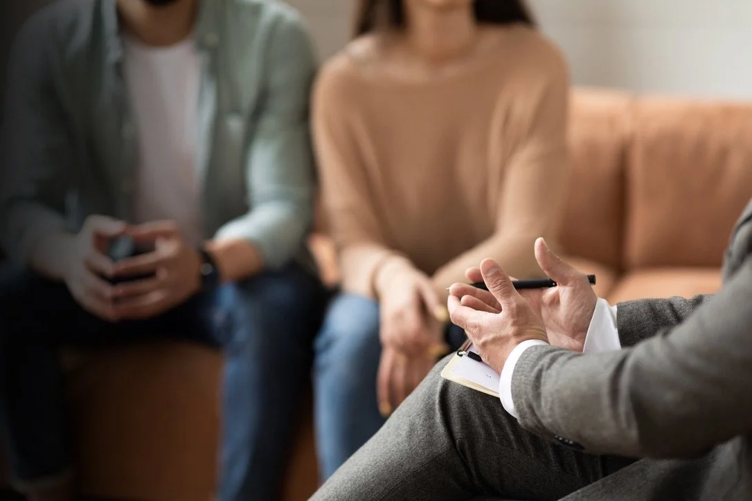 A therapist or counselor sitting with two clients during a therapy session, taking notes with a pen and notepad.