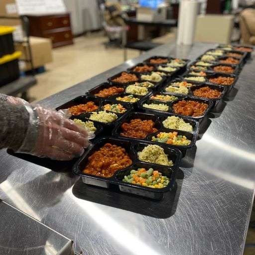 Several black food containers filled with different types of prepared meals, lined up on a stainless steel counter in a kitchen or cafeteria setting.