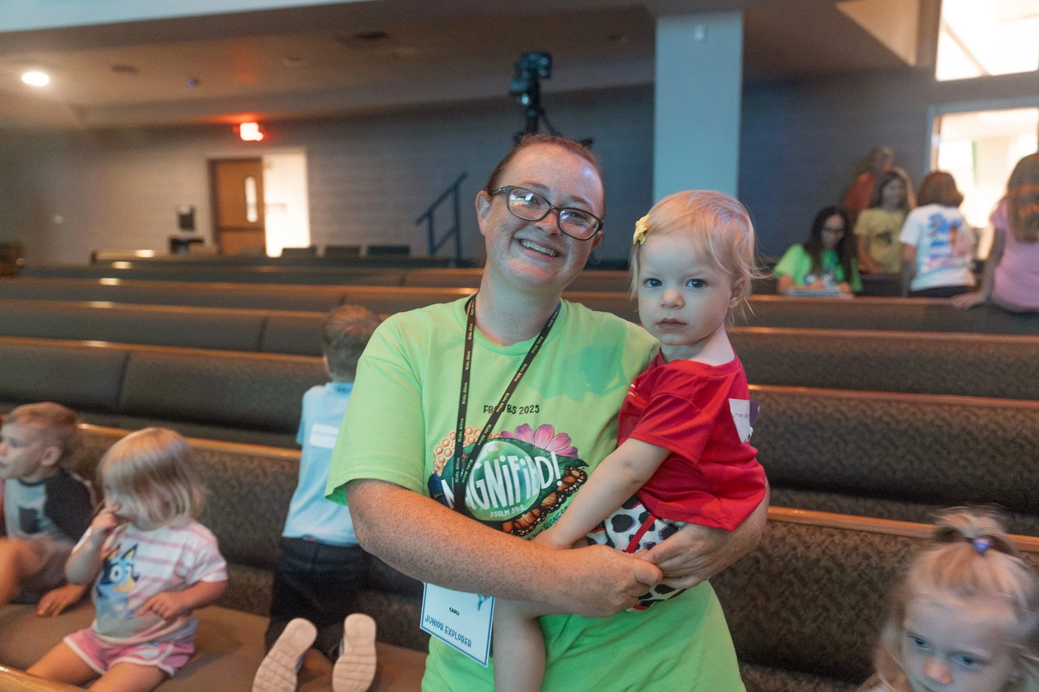 A woman with glasses and a green shirt smiling while holding a young girl with blonde hair and a red shirt in a crowded indoor auditorium.