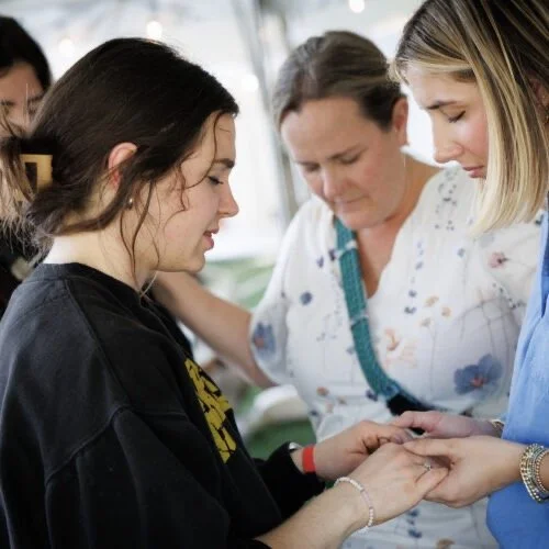 Three women in a supportive moment, with two of them gently holding hands and the third placing a hand on the other's shoulder, inside a well-lit room.