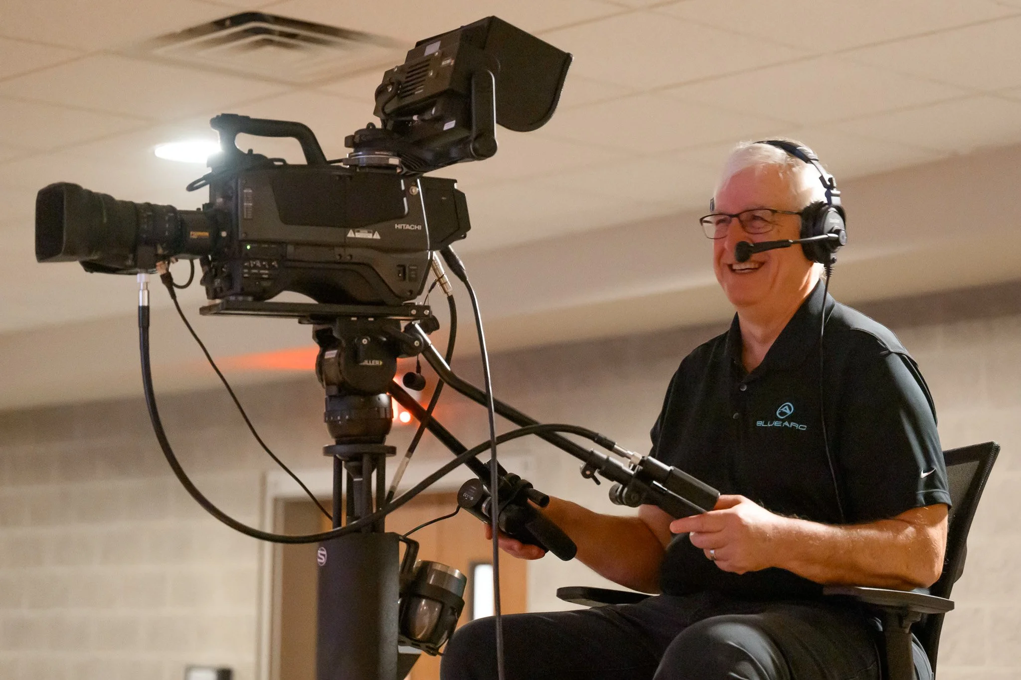 An older man with white hair and glasses operating a professional video camera. He is smiling and wearing a black polo shirt with a logo, and a headset with a microphone.