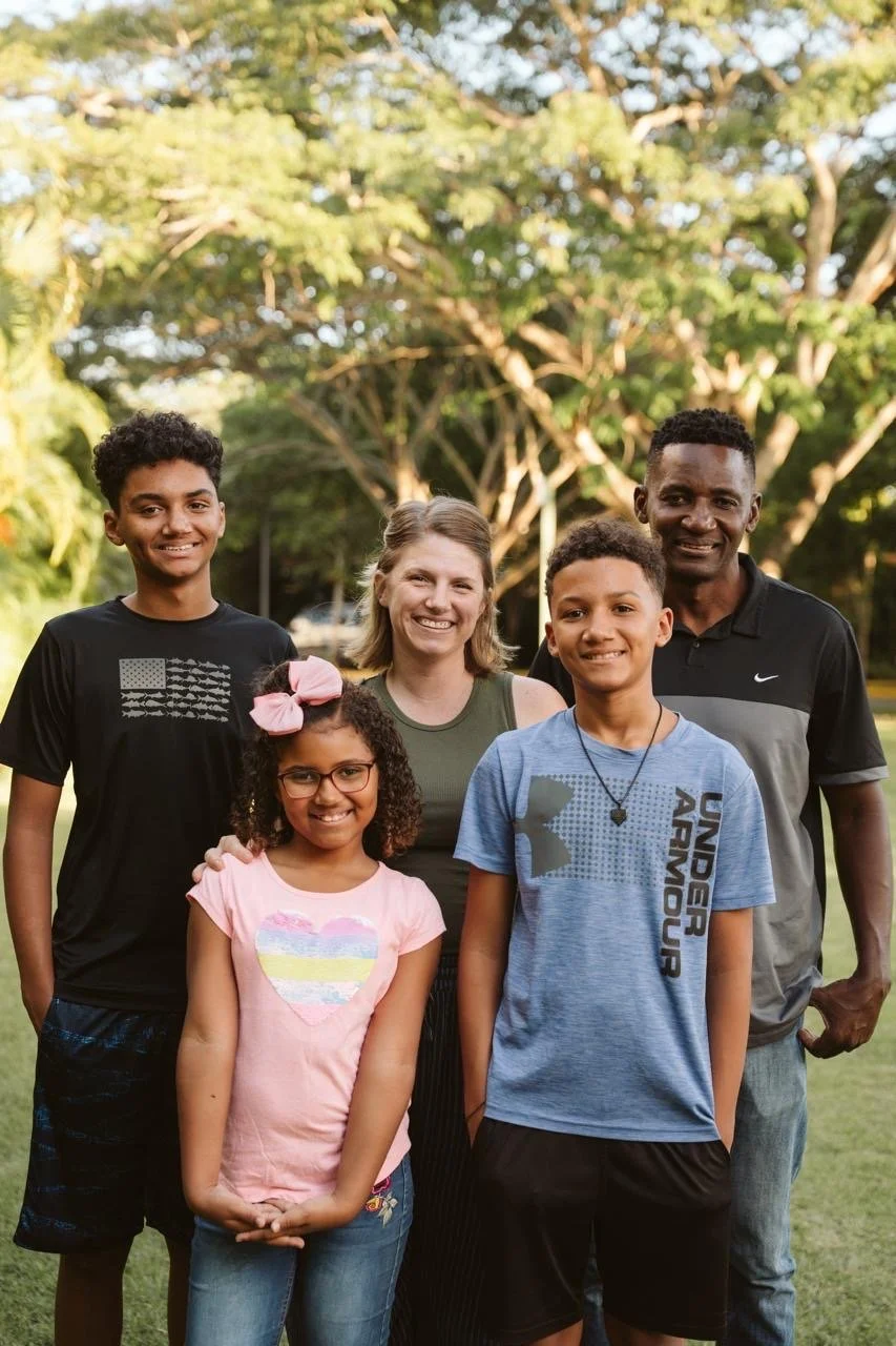 A family of six standing outdoors in a park, smiling, with trees in the background.