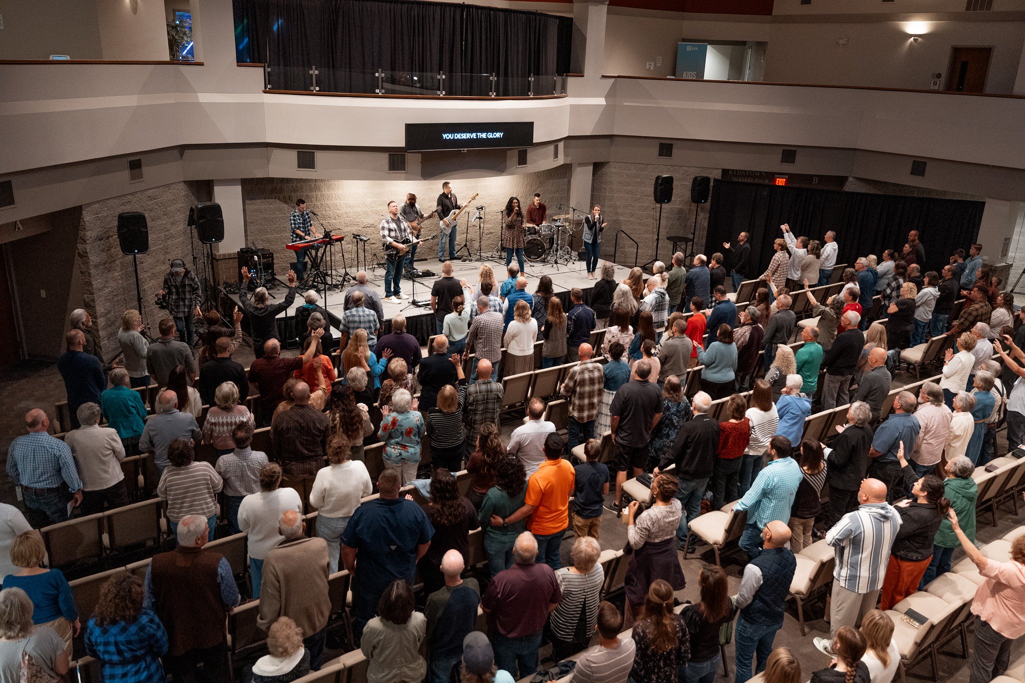 A large gathering of people attending a church service or concert in an auditorium. The stage has musicians performing and a digital screen displaying the words 'YOU DESERVE THE GLORY' above them.
