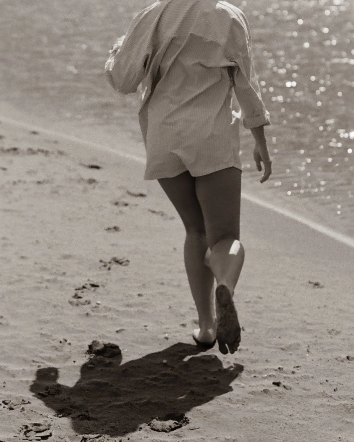 A person walking on a sandy beach, dressed in an oversized shirt, with their back to the camera. The person is barefoot and footprints are visible behind them. The water is in the background, with sunlight reflecting off the surface.