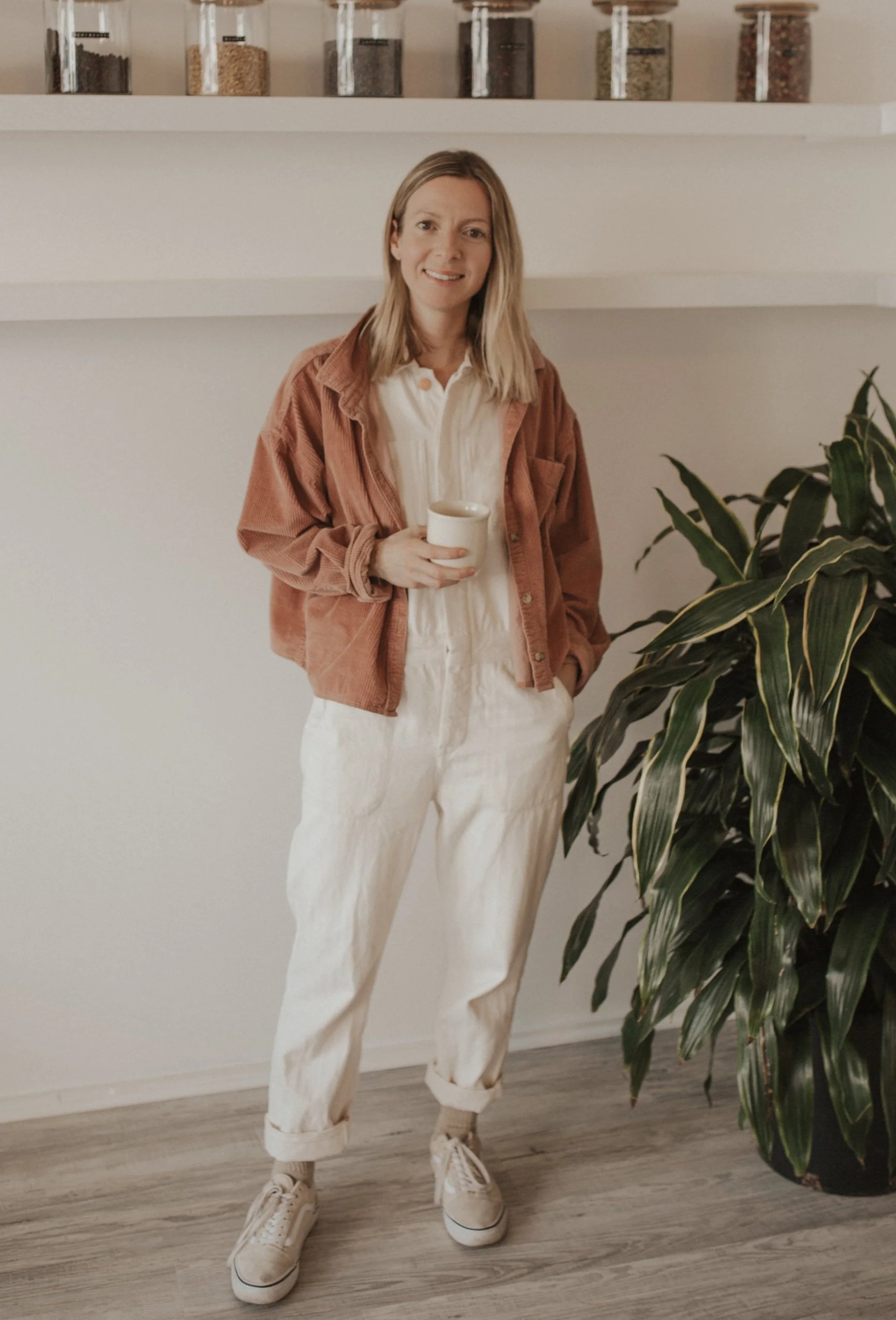 A woman with shoulder-length blonde hair wearing a white shirt, light-colored pants, a rust-colored jacket, and sneakers, standing next to a large leafy houseplant, holding a cup, indoors with shelves of spices or grains in glass jars behind her.