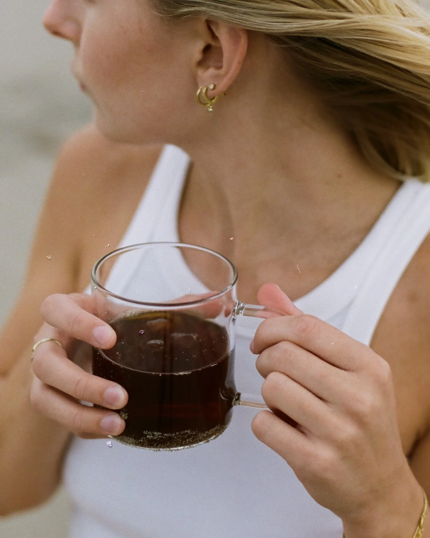 A woman with blonde hair and gold jewelry holding a glass mug filled with dark coffee, wearing a white sleeveless top.
