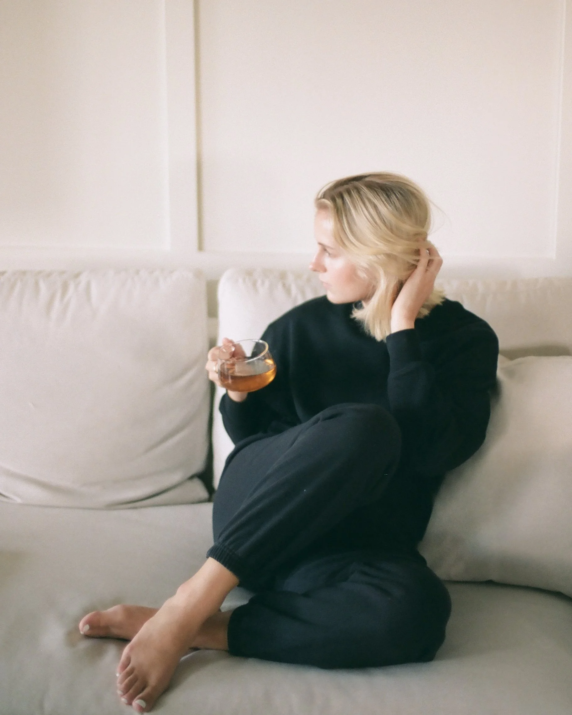Person with blonde hair sitting on the floor, holding a drink, and stretching out their legs on a beige carpet in front of a white wall with minimal decor.