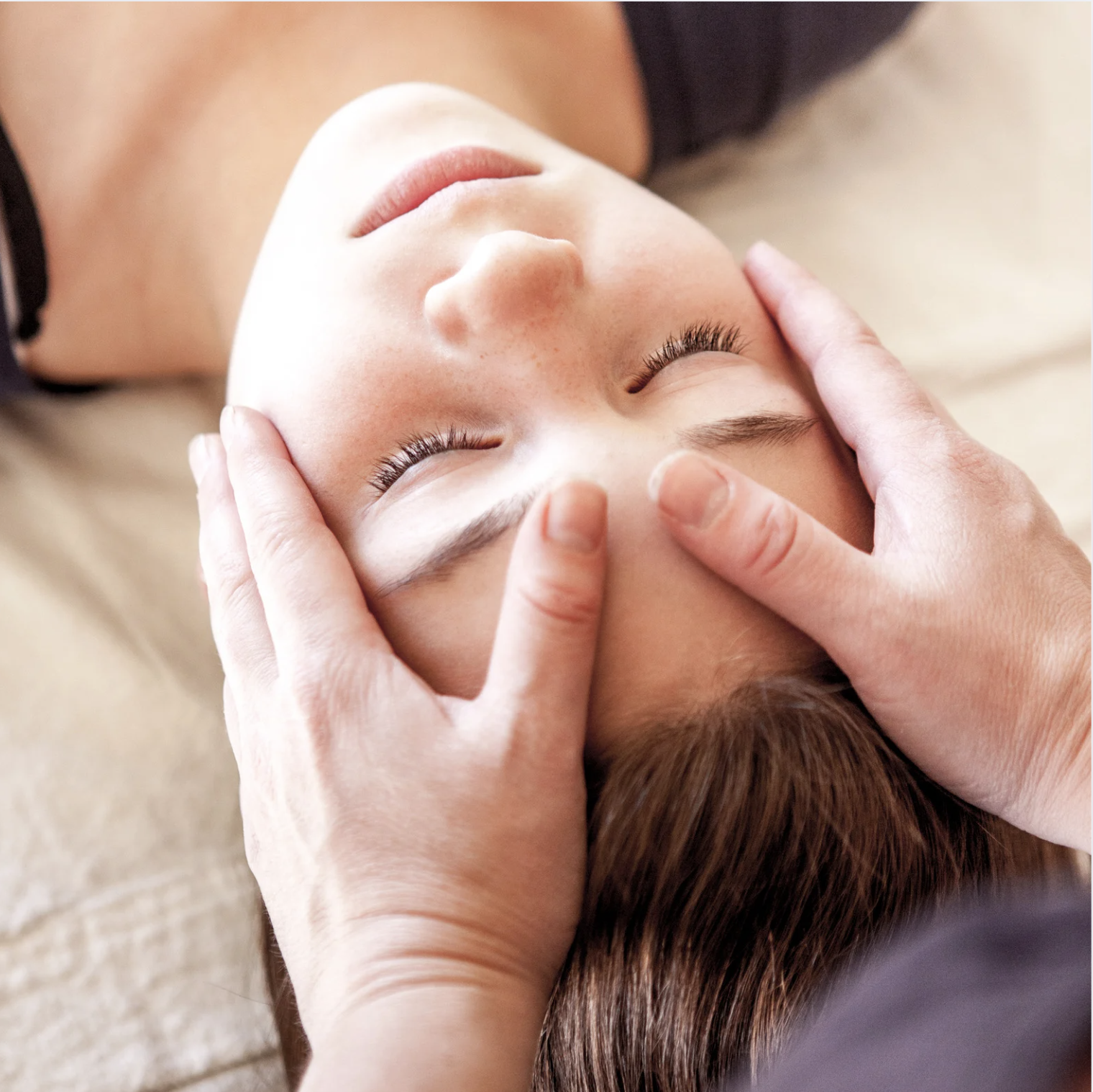 A person receiving a facial massage or treatment, lying on their back with closed eyes, while someone gently presses their temples.