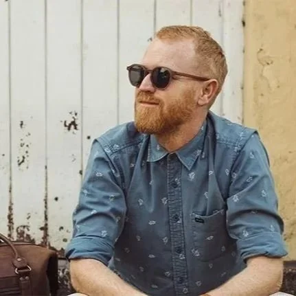 A man with red hair and beard wearing sunglasses, a blue patterned shirt, and sitting outdoors against a white wooden fence.