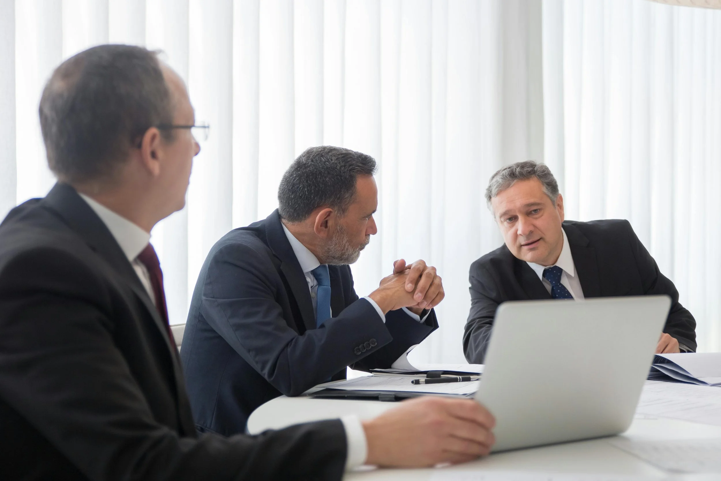 Advisory Services for the Commercial Real Estate Market. Three businessmen in formal attire having a serious discussion in a meeting room with a laptop and documents on the table.