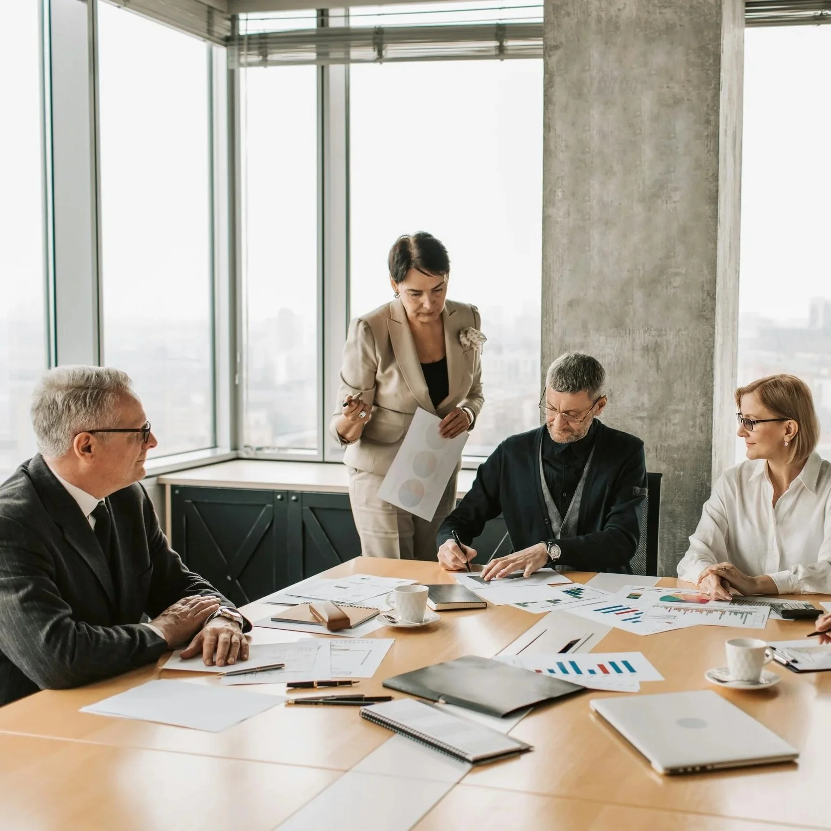 A business meeting with five people, including three men and two women, sitting around a table with papers, charts, and laptops in a modern office with large windows and a city view.