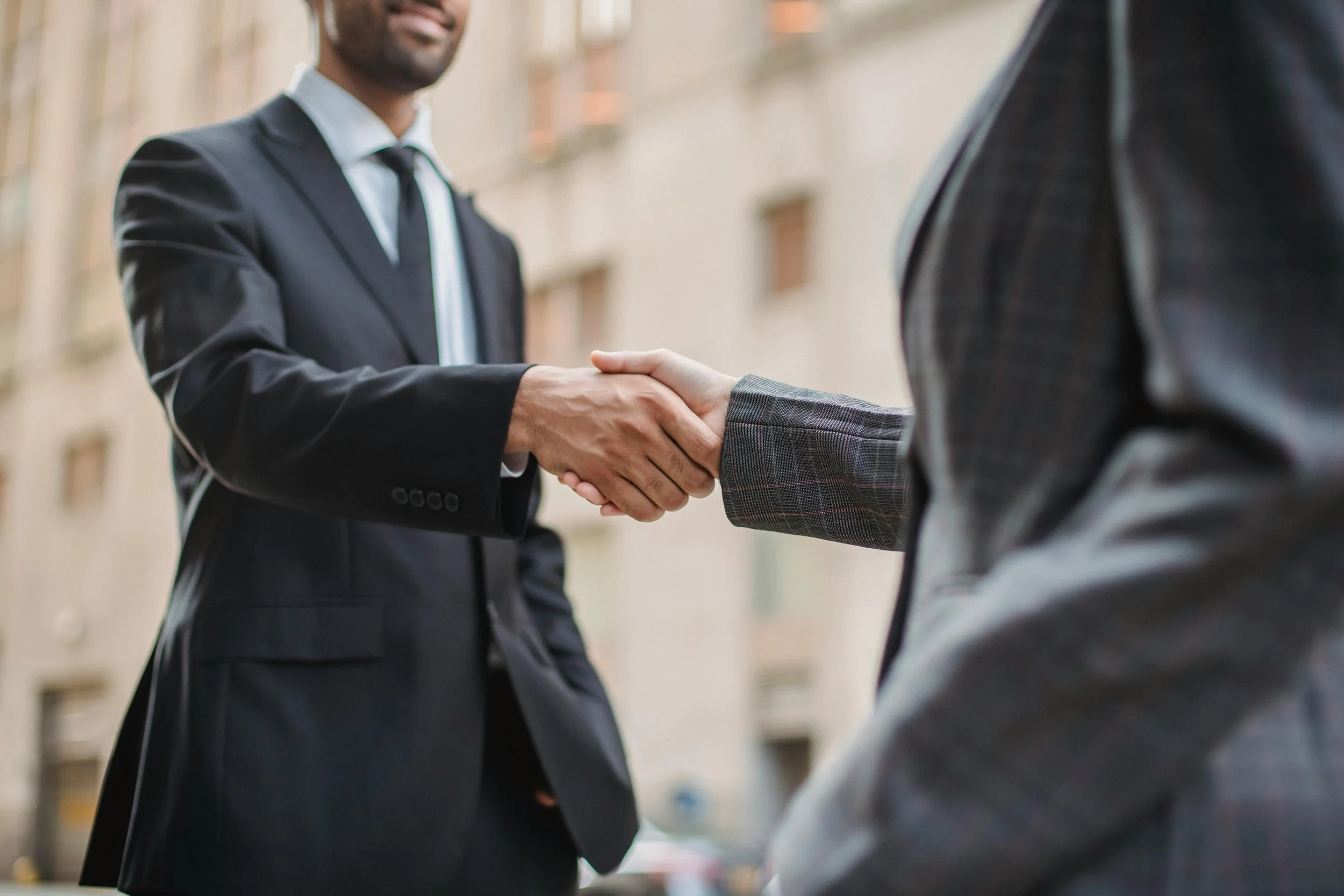 Two men in business suits shaking hands outdoors, with a blurred city building in the background.