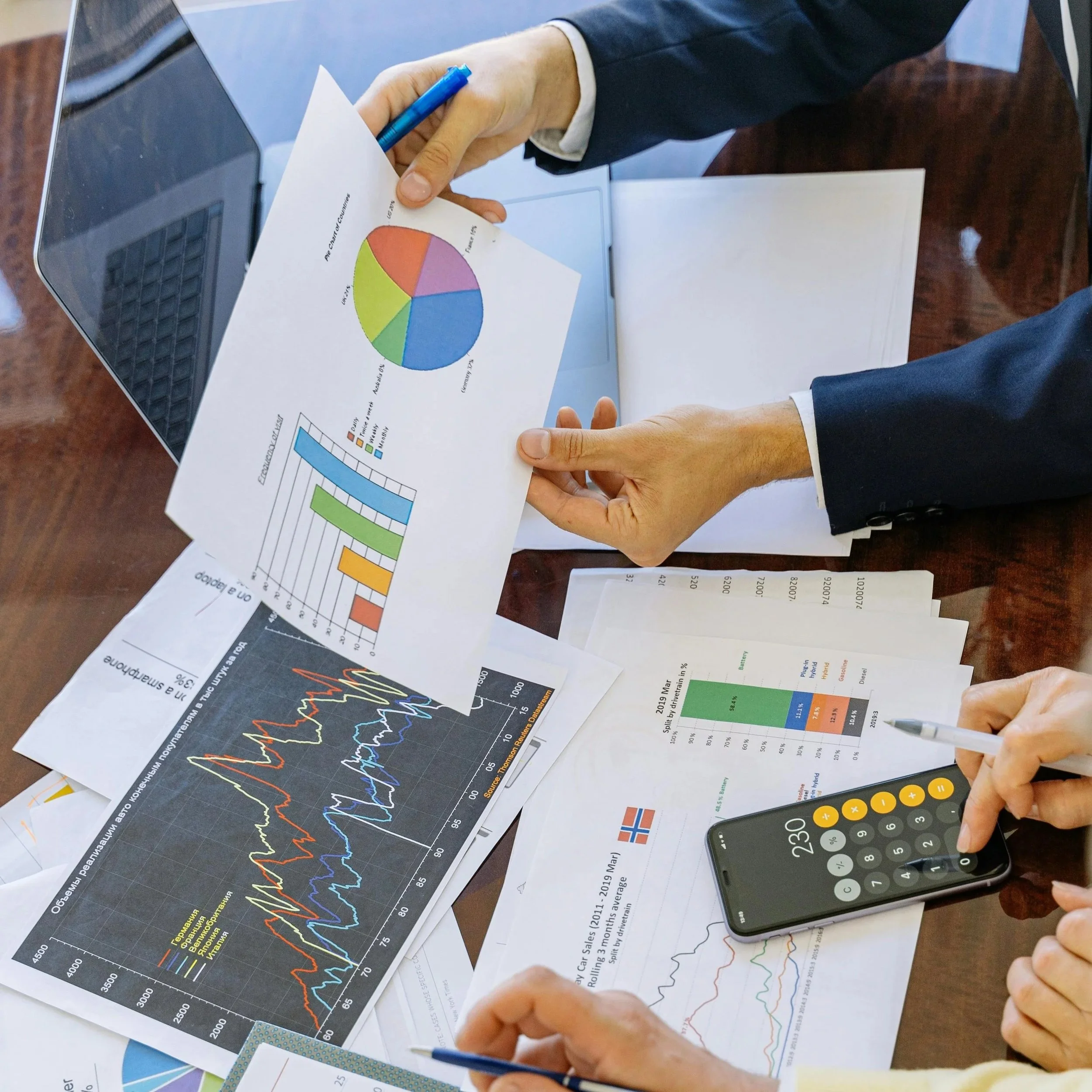 Business meeting with two people reviewing printed charts and graphs, one holding a paper with a colorful pie chart, another using a calculator, laptop, and additional documents on the table.