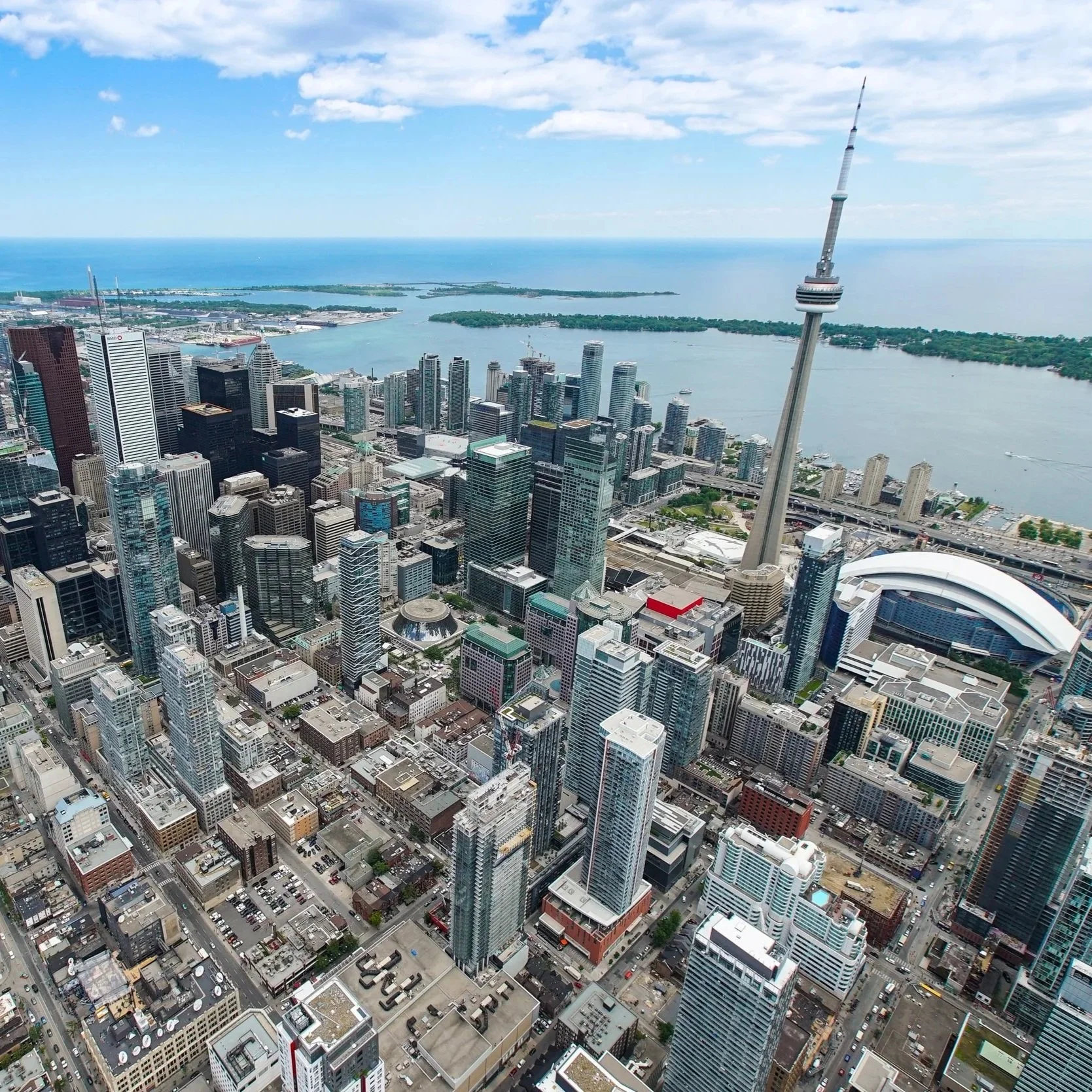 Aerial view of downtown Toronto, Canada, with the CN Tower prominently visible among many skyscrapers, near Lake Ontario on a partly cloudy day.