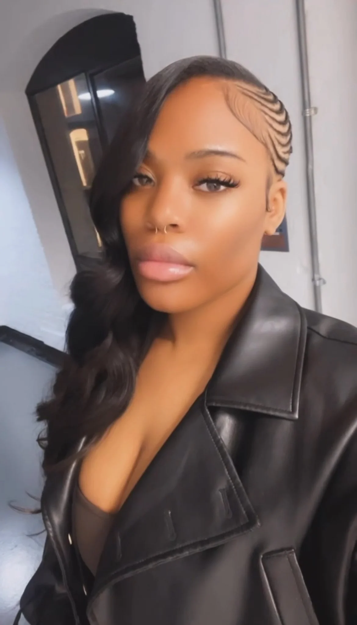 A woman with styled long wavy black hair, light makeup, and a septum piercing, wearing a black leather jacket, taking a selfie indoors.